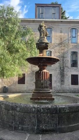 A video shows a stone fountain in a courtyard with a building in the background. The fountain is made of weathered stone and has a statue of two figures on top. Water is spraying from the fountain into a basin. The building behind the fountain is made of stone and has several windows. The sky is blue with some clouds.