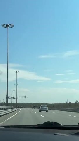 The video shows a driver's perspective while driving on a highway during the daytime. The sky is blue with some white clouds. The highway has multiple lanes with other cars moving in the same direction. There are tall light poles along the side of the road. A line of trees is visible in the distance.