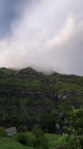 The video captures a dramatic mountain landscape under a cloudy sky. The mountains are covered in lush green vegetation with exposed rocky layers. As the video progresses, a rainbow appears, arcing across the sky to the right of the mountains. In the foreground, green trees with dense foliage are visible, and a portion of a building with a metal roof is seen on the right side of the frame. The weather appears to be clearing after a rain, as indicated by the rainbow and clouds. The scene suggests a remote, natural environment, possibly in a mountainous region.