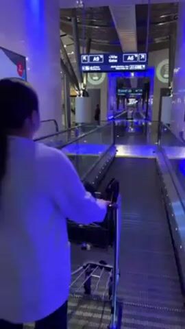 A woman in a white shirt and dark pants, with her hair in a ponytail, is pushing a luggage cart on a moving walkway in an airport. Overhead signage indicates directions to gates A9 and A6. The walkway and surrounding area are lit with blue light. Other people are seen in the distance. The airport appears modern with a reflective floor.