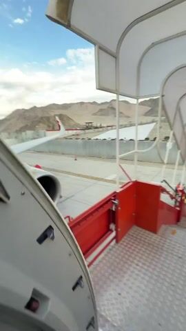The video begins with a close-up view of an airplane's red and white livery, its engine, and a set of red mobile stairs on an airport tarmac. The stairs lead up to the airplane's door. In the background, mountains are visible under a cloudy sky. The camera pans slightly, revealing more of the airplane's wing and fuselage. As the video progresses, the view shifts to looking down the red mobile stairs, where several people wearing high-visibility vests are seen walking up and down. The scene transitions to a view from the top of the stairs, looking out over the tarmac. Several people, including one in an orange vest, are visible on the ground. A line of blue and white airport vehicles is parked in the distance, and more mountains form the backdrop under the sky. The overall environment is that of an airport during daylight.