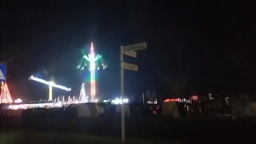 The video captures a night scene at a carnival or amusement park. In the background, several rides are illuminated with colorful lights, including a tall swing ride at the center, a crane-like ride to the left, and other structures. The rides appear to be in operation, with lights flashing and moving. In the foreground, there are traffic signs, including a blue and white pedestrian crossing sign and a red circular sign with a blue cross. Two individuals are visible, walking and looking towards the carnival. The overall atmosphere is dark, illuminated by the artificial lights of the rides and signs. The camera appears to be moving, possibly from a vehicle, as indicated by the slight blur and the angle of the shots.