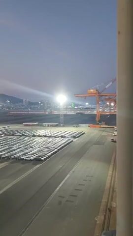 The video shows a time lapse of a large car storage lot at dusk. Cars are neatly parked in rows under bright floodlights. In the background, a city skyline and mountains are visible. A large crane is also present, suggesting the lot is near a port or industrial area. There are a few people and vehicles moving through the lot. The sky changes from a pale blue to shades of pink and orange as the sun sets.