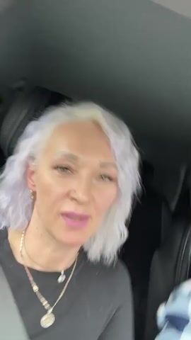 A woman with white and purple hair, wearing a black long-sleeved shirt, and two necklaces is sitting in a car. She is wearing makeup, including pink lipstick and false eyelashes. She is looking at the camera and talking. A seatbelt is visible across her chest.