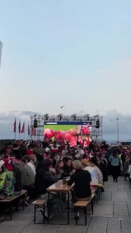The video shows a crowded outdoor gathering with people sitting at long tables, facing a large screen mounted on a scaffolding. The screen displays a live broadcast, likely a sports event, featuring several individuals. The atmosphere appears festive, with many attendees wearing red and white clothing, some with hats and scarves. Some people are holding drinks and appear to be engaged in conversation and watching the screen. The environment is an urban setting with a modern glass building to the left and flags visible in the background. Seagulls are flying in the sky, and the lighting suggests it is late afternoon or early evening. The overall impression is of a public viewing event for a significant occasion.