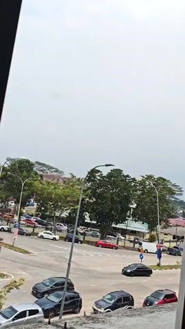 The video shows a wide shot of a traffic intersection with a roundabout. Multiple cars are visible on the road, some parked and some in motion. A lamppost stands prominently in the center of the roundabout. Trees line the background, with buildings and other structures further behind them. The sky is overcast. In the foreground, closer to the camera, there are several more cars parked or driving.