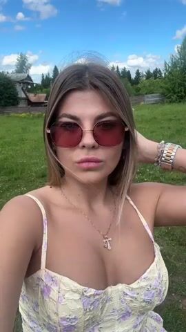 A young woman with long blonde hair, wearing a light-colored floral dress and pink-tinted sunglasses, takes a selfie outdoors on a sunny day. She is standing in a grassy area with trees and a building in the background under a blue sky with white clouds. She poses for the camera, touching her hair and sticking out her tongue.