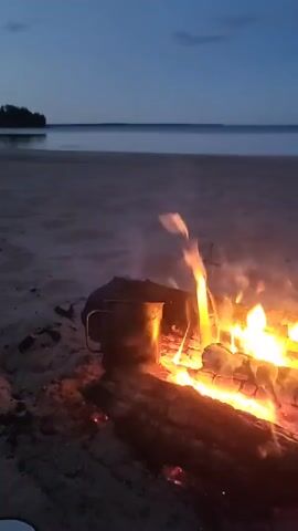 The video shows a campfire on a sandy beach at dusk. A metal mug is placed next to the fire, with steam rising from it, suggesting something is being heated. The fire burns brightly with orange and yellow flames against a backdrop of a calm body of water and a dark blue sky. The overall scene conveys a peaceful, rustic atmosphere.