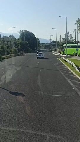 A wide-angle video shot from the perspective of someone in a vehicle on a road in Neos Panteleimonas, Greece. The road is paved and appears to be a highway with multiple lanes. The sky is clear and blue, suggesting it is daytime, likely late morning or early afternoon, with bright sunlight casting shadows.

In the foreground, the road surface is visible, with lane markings clearly delineated. There is a reflection on the windshield, showing a blurred image of what appears to be a person wearing sunglasses and a hat, and possibly part of the vehicle's interior.

Further down the road, a white car is driving in the same direction as the camera's vehicle. To the right of the road, there is a green bus with the number "50" visible on its side. A metal guardrail runs along the side of the road, separating it from the surrounding landscape.

The landscape consists of lush green trees and vegetation, with some hills or mountains visible in the distance. There are several tall lampposts lining the road, as well as utility poles with wires. The scene conveys a sense of travel and a sunny day in a rural or semi-rural Greek setting.

The audio consists of a voice speaking in Russian, discussing ancient Greek beliefs about the creation of the first humans by the gods. The timestamp in the audio indicates "00:00 - 00:04", suggesting the beginning of this narrative.