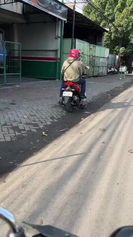 A person wearing a pink helmet and a tan hoodie rides a red scooter away from the camera on a paved road. The scooter has a white license plate and a black seat. The person is wearing dark pants. The road is lined with trees and buildings. The sun is casting long shadows on the ground. The video appears to be taken from another vehicle.
