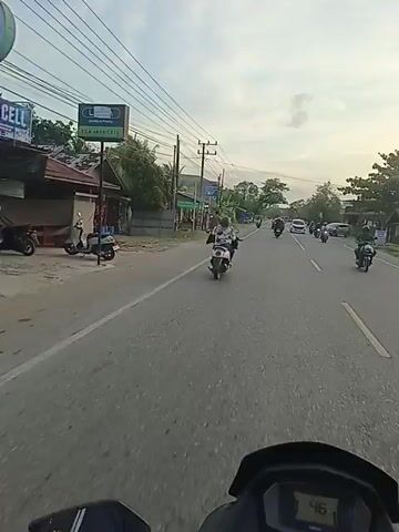 The video shows a view from a moving motorcycle on a paved road. The road has white lines marking lanes.  There are other vehicles on the road, including cars and motorcycles.  A person walks along the side of the road. There are buildings, trees, and utility poles visible along the roadside.  The sky is overcast.