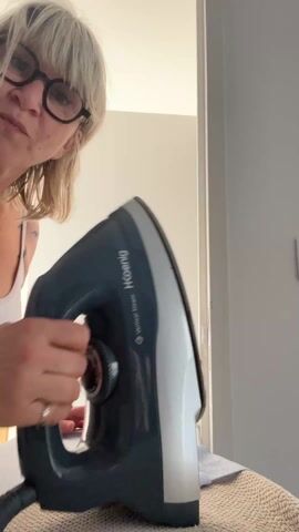 A woman with short blonde hair and glasses is shown from the chest up, facing the camera. She is holding a H.Koenig iron, showing its underside before activating the steam function by pressing a button. The iron is grey and white with a silver soleplate. An ironing board and a partially ironed blue and white striped garment are visible below the iron. The background includes a white wall and a door.