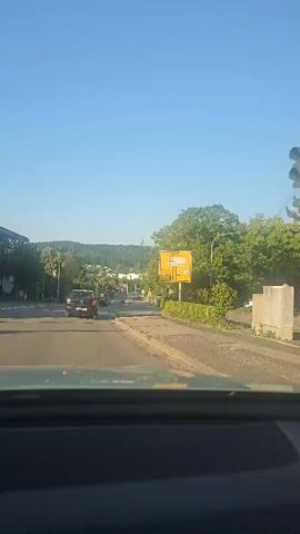 The video shows a view from a car driving down a road on a clear day. On the right side of the road, there is a yellow sign with text and arrows indicating directions to various locations. Further down the road, trees line the right side, and on the left, there are buildings and trees. Several cars are visible on the road, including one black car ahead of the vehicle from which the video is being recorded. In the distance, there are hills with buildings and a structure that appears to be a tower. The sky is clear and blue.