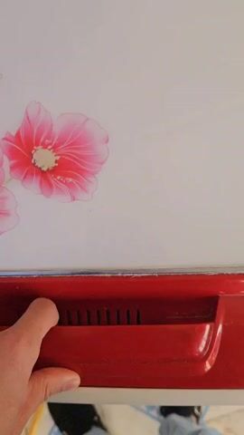A hand opens the top of a red and white portable washing machine, revealing sudsy water inside. A folded off-white cloth is then placed into the water.