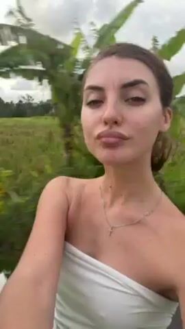 The video shows a woman sitting in a moving vehicle, possibly a golf cart, with rice fields visible in the background. She is wearing a white strapless top and a delicate necklace.  She waves and poses for the camera, occasionally running her hand through her hair. A green and black object is visible next to her, possibly a helmet or bag. The sky is overcast.