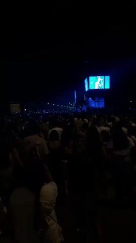 A dark video shows a crowded outdoor event at night. There is a large screen in the background displaying a person. People are standing and milling around, with some appearing to be dancing. The lighting is dim, with some blue stage lights visible in the distance.