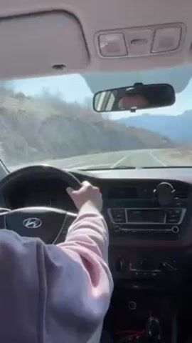 The camera is situated inside a car, capturing a driver's perspective of the road ahead. A person is seen holding the steering wheel, wearing a light pink sweatshirt. The interior of the car features a dark brown dashboard with a standard car radio and air conditioning controls. The car's logo, Hyundai, is visible on the steering wheel. Outside, the road winds through a mountainous region under a clear, blue sky. The mountains are covered with snow-capped peaks, and the roadside is lined with sparse trees. The rearview mirror shows the reflection of the driver's face, partially obscured by sunglasses. The car appears to be moving smoothly along the road.