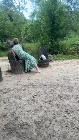This video shows a person sitting on a carved wooden bench in a park-like setting. The bench is carved to resemble a person lounging. The person, wearing a green and white patterned dress and white sneakers, is leaning back on the bench, looking away from the camera. A light-colored purse is placed on the bench next to them.

The scene is an outdoor area with sandy ground, typical of a path or clearing in a wooded area. In the background, there are several other carved wooden seating structures, some resembling chairs and others more abstract shapes. Behind the seating, lush green trees and foliage create a dense woodland backdrop. The lighting suggests it is daytime, likely late morning or afternoon, with soft, diffused sunlight filtering through the trees, indicating potentially overcast or partly cloudy weather.

The overall impression is one of a peaceful, natural environment. The person appears relaxed, enjoying a moment of rest in a tranquil setting. There is no visible text in the video. The location context of Gravesham, United Kingdom, suggests this could be a public park or a designated nature area within that region, possibly featuring art installations like the carved benches.