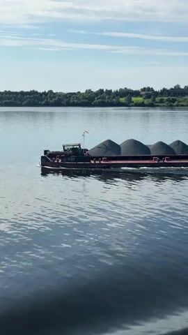 The video shows a barge laden with gravel being towed by a tugboat on a wide river. The barge is long and flat, with several evenly spaced mounds of dark gray gravel filling its cargo area. A small orange flag flies from the front of the barge. The tugboat is white and has a taller superstructure with windows. It is positioned behind the barge, connected by a tow line, and is actively pulling it forward. The water is calm with gentle ripples, reflecting the blue sky and scattered white clouds. The riverbanks in the background are lined with dense green trees and a few scattered buildings, including what appears to be a red brick tower with a dome. The scene is illuminated by bright daylight, suggesting a clear and pleasant weather day. The camera is positioned on the water, moving alongside the tugboat and barge, offering a view from the side and slightly ahead of the procession.