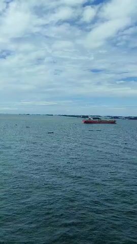 The video shows a wide shot of a large body of water under a cloudy sky. A red cargo ship is visible in the distance, moving across the water. Several other dark-colored objects, possibly other ships or structures, are also scattered across the horizon. The water is choppy with visible waves. The sky is partly cloudy with white clouds against a blue background. The camera appears to be panning slightly to the right.