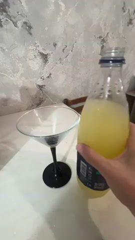 A person's hand holds a bottle containing a light-yellow liquid, pouring it into a clear martini glass with a black stem and base. The glass is positioned on a white surface, which may be a table. In the background, a wall with a textured, marbled gray pattern is visible, and a wooden chair or furniture piece is partially seen. The liquid in the bottle appears to be a beverage, possibly lemonade or a cocktail mix. The pouring action is captured from a slightly overhead angle.