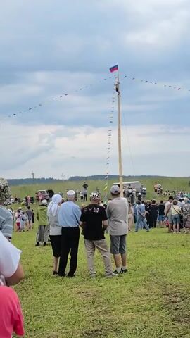 A group of people are gathered in a grassy field, watching a man climb a tall wooden pole. The man is near the top of the pole, where a Russian flag is flying. The pole is decorated with a string of colorful flags. The sky is cloudy. Several vehicles, including a white trailer, are visible in the background, as well as more spectators.