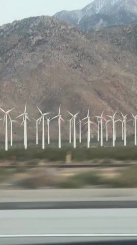 The video captures a wide shot of a large wind farm from a moving vehicle. In the foreground, a blurred highway guardrail and road indicate motion. Parallel to the road are train tracks. Beyond these, numerous white wind turbines stand tall against a backdrop of arid, rocky mountains. Many of the turbines have small red lights on their nacelles. The blades of most turbines appear stationary or are moving very slowly. The mountains in the background have patches of snow on their highest peaks. The sky is clear, suggesting daytime. The scene evokes a sense of vastness and renewable energy infrastructure in a desert environment, consistent with the location context of Cabazon, United States. There are no people visible, and no text is present.