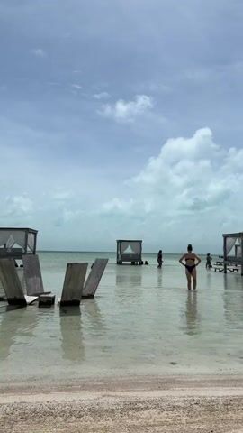 A woman in a dark bikini stands in shallow water on a beach under a cloudy sky. Several people are in the water in the background, near small cabanas built on stilts. Three wooden chaise lounges are partially submerged in the water in the foreground, near the sandy shore.