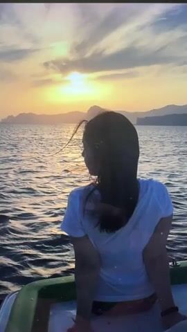 A person, viewed from behind, sits on a boat, looking out at a sunset or sunrise over a vast body of water. The person has dark, long hair, with some strands appearing to be gently blown by the wind, and is wearing a light-colored t-shirt. The sun, a bright golden orb, is low on the horizon, casting a warm glow across the sky, which features scattered clouds. Its light reflects on the rippling surface of the water. In the distance, dark silhouettes of mountains or cliffs are visible along the coastline. The foreground shows a partial view of the boat, with a distinctive green edge. The scene evokes a sense of peace and contemplation amidst natural beauty. There is no visible text.
