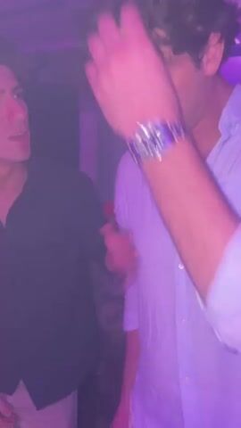 The video depicts a dimly lit indoor scene, likely a party or club, with predominantly purple and blue lighting. Two men are in the foreground. The man on the left, wearing a dark shirt and light-colored shorts, is looking towards the right and gesturing with his left hand. The man on the right, wearing a light-colored collared shirt, has his right hand raised to his head. In the background, a DJ booth with illuminated controls is visible. Further in the background, a crowd of people can be seen dancing and socializing, illuminated by shifting colored lights, including red and blue. The overall atmosphere is energetic and social.