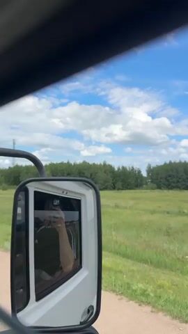 The video is taken from inside a vehicle, showing the side mirror reflecting the interior and the person holding the phone. The exterior view shows a dirt road, a grassy field, a line of trees, and a blue sky with white clouds. A tall tower, possibly a communication tower, is visible in the distance. The vehicle appears to be moving, as indicated by the blur of the passing scenery.