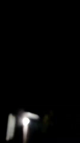 A short video clip captures a nighttime scene. The video begins with a completely black screen, followed by a blurry light that briefly appears at the bottom left. Then, a distant, purplish glow illuminates the sky, suggesting lightning or a very bright light source in the distance. The sky is filled with dark, indistinct clouds. Silhouettes of trees and possibly buildings are visible against the illuminated sky. The video then cuts back to a black screen.