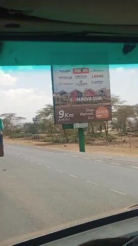 The video shows a view from the inside of a vehicle driving on a paved highway in a rural area. A large billboard advertising Naivasha, located 9km ahead, is visible on the right side of the road. The environment features dry grassland and scattered trees. The camera pans slightly, focusing on the road ahead, where a truck with a blue tarp-covered load is traveling in the same direction. Another truck is visible in the distance, approaching from the opposite direction. The dashboard of the vehicle is visible, with a sticker and wiper blades in view. The sky is partly cloudy.