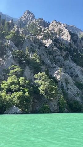 The video captures a scenic natural landscape under a clear blue sky. A large, rugged mountain range, composed primarily of grey rock, dominates the background. The steep mountain slopes are sparsely covered with green foliage, including trees and bushes, clinging to the rock faces.

In the foreground, a vibrant, turquoise-colored body of water fills the lower portion of the frame, showing gentle ripples on its surface. The water's distinct color suggests mineral content typical of certain rivers or dam lakes in mountainous regions.

The video appears to be a static shot, likely recorded from a boat or a fixed position overlooking the water, with slight movements indicating it might be handheld. The setting is a bright, sunny day, with clear skies. No people or visible text are present in the video. The natural environment suggests a canyon or gorge, aligning with the provided location context of Akseki, Turkey. Background music is audible throughout the clip.