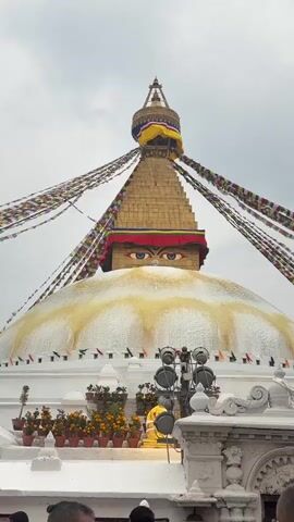 The video shows a close-up of the Boudhanath Stupa in Kathmandu, Nepal. The iconic structure is a large, white dome topped with a golden spire decorated with painted eyes. Prayer flags are draped around the stupa, fluttering in the breeze. The sky is overcast. People are visible at the bottom of the frame, suggesting the area is a popular gathering place. There are potted plants lined along a ledge at the base of the dome. The video is shot from a low angle, emphasizing the size and grandeur of the stupa.