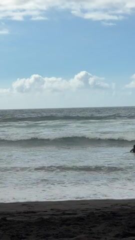The video depicts a scenic ocean view with waves rolling onto a dark sandy beach. The sky above is a vibrant blue, dotted with fluffy white clouds, suggesting a pleasant, likely daytime, weather condition. The ocean itself is a deep gray-blue, with several rows of waves visible, each breaking and forming white foam as it approaches the shore. The foreground is dominated by the dark sand of the beach, providing a stark contrast to the lighter tones of the sea and sky. The overall impression is one of a tranquil yet dynamic coastal environment. There is a person visible in the water on the right side of the frame, appearing to be swimming or playing in the waves, adding a subtle human element to the natural landscape. The location is identified as Puerto de la Cruz, Spain. There is no visible text in the media.