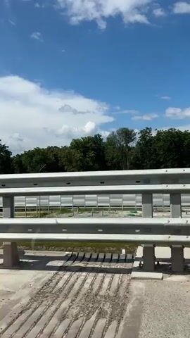 The video shows a view from a vehicle moving along a road, likely a bridge. In the foreground, there is a concrete barrier with a metal guardrail above it. The guardrail is made of multiple horizontal metal bars, supported by vertical metal posts. To the left of the guardrail, there's a small red reflector. The road surface appears to be concrete or asphalt, with a drain cover visible on the far left. Beyond the guardrail, a body of water, possibly a river or lake, is visible. There's a sandy beach area along the water's edge, with some sparse vegetation. Further in the distance, a treeline of lush green trees forms the horizon. Above, a bright blue sky is dotted with fluffy white clouds, some of which appear to be cumulus clouds. A faint white contrail from an aircraft is visible in the sky. The overall impression is of a scenic drive on a sunny day, possibly during the summer. The movement of the vehicle causes the scenery to shift and the perspective to change slightly throughout the short clip. No people are visible in the video.