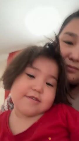 A young child with dark hair and a red shirt smiles and makes a peace sign. An adult with dark hair is in the background. The child is in focus, and the adult is slightly out of focus. The background is blurred, but appears to be an indoor setting with a light-colored wall and a piece of furniture. The video is shaky and appears to be a selfie.