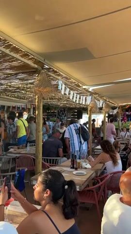 This is a vertical, eye-level, medium shot of an outdoor dining area under a large, beige awning. The setting appears to be a restaurant or bar, possibly near a beach given the light and open-air feel. Numerous people are seated at wooden tables with chairs. The tables are cluttered with plates of food, drinks, and napkins. Many patrons are wearing light-colored clothing, and several individuals are wearing blue and white striped jerseys, suggesting a sporting event or fan gathering. In the background, a large screen displays an image of a man, likely a sports figure, with text above him. Some people are actively engaged in conversation, while others are looking at their phones or food. The overall atmosphere seems casual and lively, with a moderate number of people spread throughout the space. The lighting suggests it is daytime, possibly late afternoon.