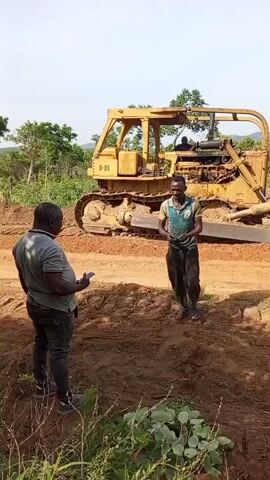 In a daytime scene in Otunade, Nigeria, two men stand on a dirt path with a large yellow bulldozer in the background. The man on the left, wearing a grey t-shirt and dark pants, holds a mobile phone while looking at the man on the right. The man on the right, dressed in a patterned blue and green t-shirt and dark pants, is holding small items in his hands, appearing to show them to the other man.

The bulldozer, marked "D-8K" on its side, is parked on the cleared earth, suggesting a construction or land development site. Lush green trees and foliage frame the background, indicating a rural or undeveloped setting. The sky is clear, and the overall lighting suggests a sunny day. The men appear calm and engaged in their conversation.