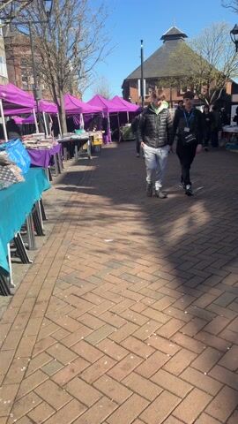 The video opens with a view down a bustling outdoor market walkway on a clear, sunny day. The ground is paved with reddish-brown brick pavers, and long shadows are cast by unseen objects, indicating a relatively high sun.

On the left side of the walkway, a line of market stalls with vibrant purple canopies are set up. Each stall has tables covered with matching purple tablecloths, displaying various folded goods that appear to be textiles, fabrics, or clothing items. People can be seen browsing or attending the stalls further down the line.

Initially, two young men walk towards the camera and then past it, moving away from the viewer. One wears a black jacket, light grey trousers, and white trainers, with a lanyard around his neck. The other is in a black jacket, dark trousers, and dark shoes, also wearing a lanyard.

In the background, a distinctive brick building with a dark, possibly conical or domed, roof stands out against the blue sky. Other less defined buildings are also visible. Bare-branched trees line parts of the walkway, suggesting it might be early spring.

As the video progresses, the camera pans slightly to the left, offering a closer view of the market stalls and their displayed merchandise. It then pans back to the right, showing more of the walkway and the people walking in the distance towards the conical-roofed building. Throughout the video, the market appears active but not overly crowded, with a relaxed atmosphere under the bright sunshine. Some small pieces of litter are visible on the paved ground.