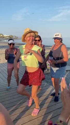 At the 00:00 mark, a group of four women are on a wooden boardwalk near a beach. The woman in the foreground, wearing a straw hat and a yellow cropped shirt with a red leopard print skirt, is in the center of the frame and appears to be dancing. The woman to her left, wearing a blue tank top and pink shorts, is clapping her hands. The woman to the right, wearing a grey tank top and a blue skirt, is holding a cup. Another woman, wearing a purple patterned top, is standing behind the dancer. In the background, the ocean, beach, and some buildings are visible under a clear blue sky. As the video progresses, the woman in the center continues to dance, with her friends watching and cheering her on. The woman on the right takes a sip from her cup at 00:02. The scene appears to be a casual social gathering outdoors.