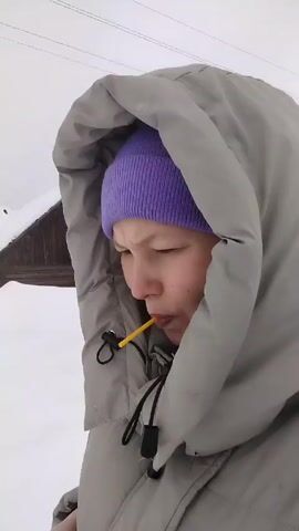 A woman in a purple beanie and gray hooded winter coat stands outdoors in a snowy environment. She has a yellow stick, possibly a lollipop or straw, in her mouth. She looks around, her expression serious. In the background, there's a small wooden structure covered in snow and power lines against a cloudy sky.