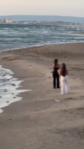 Two young women are on a sandy beach. The woman on the left wears a black top and black flared pants, posing with her hands near her head. The woman on the right wears a white top and white flared pants, holding a smartphone pointed towards the first woman, seemingly taking a photo or video. Her hair is blowing in the wind. Waves crash on the shore to the left, and a city skyline and pier are visible in the distant background.