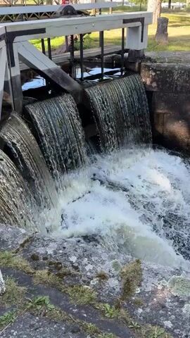 A video shows a close-up view of water flowing over a spillway. The water is white and foamy as it cascades down, creating a small waterfall. The spillway appears to be made of stone and concrete, with some moss growing on the edges. In the background, a wooden structure with white accents can be seen, possibly part of a lock or dam system. The environment appears to be outdoors, with some greenery visible. The video is short and focused on the movement of the water.