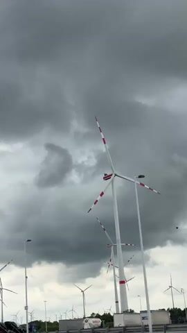 The video shows a row of wind turbines against a cloudy sky. The turbines are white with red stripes at the end of the blades. The sky is overcast with dark, ominous clouds. In the foreground, there are light poles and a highway with parked trucks. The wind turbines are spinning, indicating a windy day.