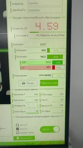 The video shows a close-up of a computer monitor displaying a manufacturing control interface. The interface shows various statistics including operator information, serial numbers, speed, and counts of items. There are also visual representations of items on the screen, some appearing to be capsules or pill-like objects. The screen displays data such as "КОЛИЧЕСТВО" (Quantity), "СКОРОСТЬ" (Speed), and other numerical values and percentages. In the background, out of focus, are industrial equipment components, possibly part of a production line, and a white wall. The lighting appears to be standard indoor lighting. The camera is held by a person, as evidenced by slight camera shake and movement. The scene is likely an industrial or pharmaceutical manufacturing environment.