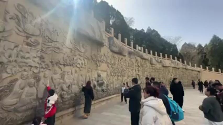 A sunny outdoor shot shows a crowd of people viewing a long, carved stone wall. The wall features detailed relief carvings of people and mythical figures. Many viewers are taking photos or videos of the wall with their mobile devices. The wall is topped with a low balustrade. Behind the wall, a line of trees borders the area. The sky is mostly clear with a bright sun shining. The ground is paved with light-colored stones.