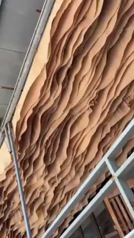 The camera points up at a ceiling-like structure constructed of layers of curved, brown material, perhaps wood or fabric, creating a wavelike pattern. A white metal frame is visible along the edges and bottom of the frame, suggesting a construction or support element. The lighting is bright, and the shot is static with no camera movement beyond handheld shakiness.