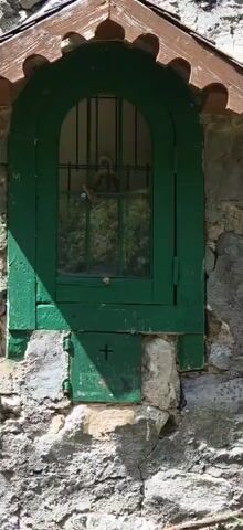 The camera pans out from a close-up shot of a small, green-framed window set into a stone wall. The window has a small, decorative roof.  The camera pulls back to show that the window is part of a one-story building made of rough-hewn stones. The building has two doorways, one with a green door and the other without a door. There is green grass around the building, and a forested hill in the background, under a clear, sunny sky.